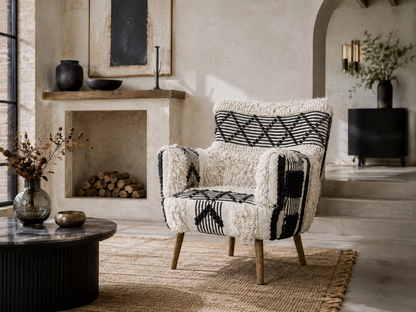 A boho armchair upholstered in a textured black and ivory patterned fabric with a fluffy cream sherpa style backrest, set in a styled living room next to a fireplace.